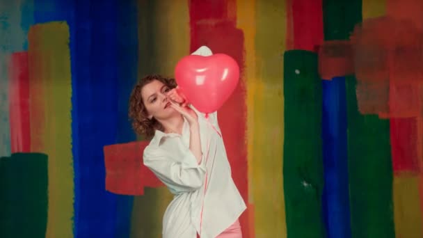 Chica atractiva con el pelo rubio rizado sosteniendo globos de aire rojo posando en la cámara. Mujer divertida con un globo rojo en forma de corazón en sus manos . — Vídeos de Stock