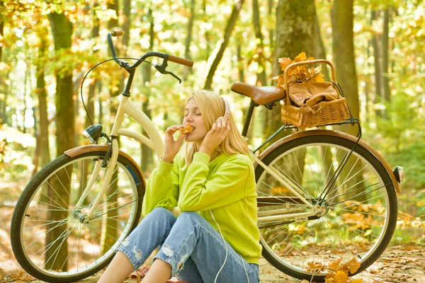 Schöne Herbst Frau beim Picknick im Herbst Wald sitzt auf dem Gras und isst. Herbstspaziergang mit dem Fahrrad. Radfahren und aktiver Lebensstil. Blonde Frau hört Musik über Kopfhörer. — Stockfoto