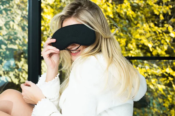 Mujer alegre con una máscara de sueño esponjosa ríe. Mujer escalofriante en máscara ocular y pijama de seda riendo en casa . — Foto de Stock