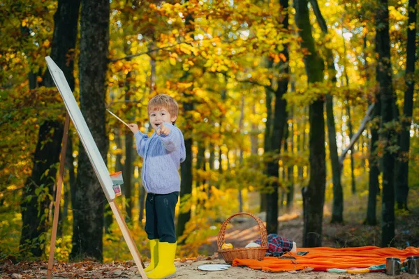 가을 숲 공원 배경에 이젤 그림을 그리고 웃는 어린 소년. 아이들 이 카메라를 가리키고 있습니다. 재밌는 행복 한 꼬마 야. 어린이 창의력의 개념. — 스톡 사진