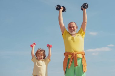 Büyükbaba ve çocuk spor salonunda ağırlık çalışıyor. Sağlıklı bir baba-oğul portresi mavi gökyüzü arka planında dambıllarla çalışıyor..