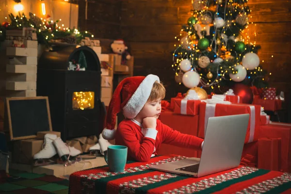 Lista de deseos para Santa Claus. Carta de tipo niño a Santa. Compras de Navidad en línea para niños. El niño está usando ropa de Santa sentado junto a su portátil. Concepto de Navidad . —  Fotos de Stock