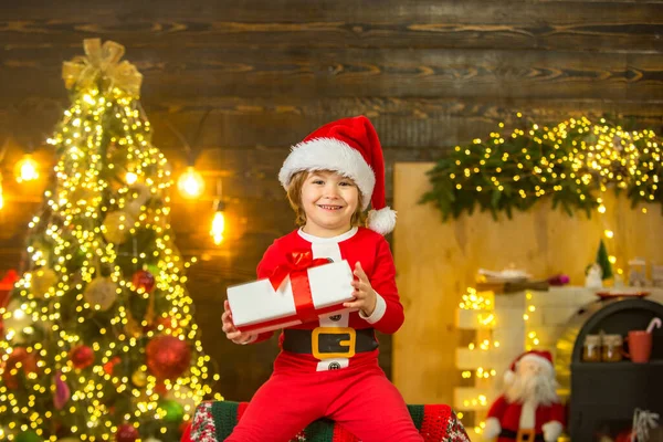 Porträtt av tomten unge med gåva tittar på kameran. Ungen har kul nära julgran inomhus. Julbarn - lycka koncept. — Stockfoto