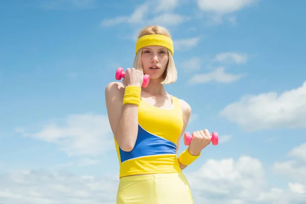 Joven y saludable deportista atractiva vestida de amarillo con uniforme de entrenamiento azul demostrando ejercicios con mancuernas rosadas en el fondo del cielo con nubes. Moda y tendencias en el deporte. Salud y —  Fotos de Stock