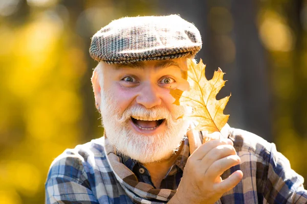 Szczęśliwy dziadek trzyma żółty liść na jesiennym tle liści. Dziadek dobrze się bawi w jesiennym parku. Portret starszego mężczyzny w jesiennym parku. — Zdjęcie stockowe