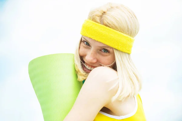 Young active and attractive woman in yellow headband holding yoga mat. Happy smiling woman going to do her yoga exercises. Sport activities, health and wellness concept.