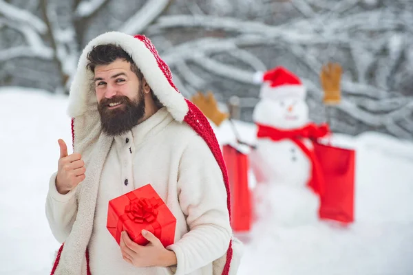 Vacances d'hiver et concept de personnes. Bonhomme de neige et homme barbu drôle avec un cadeau - les amis. Père heureux avec cadeau de Noël sur fond de bonhomme de neige d'hiver . — Photo