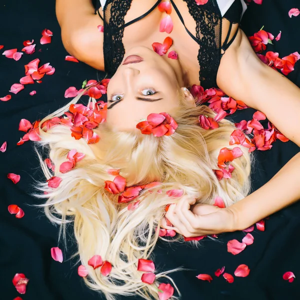 Top view of young woman in bed. Fashionable blonde beauty among falling rose petals. Woman body with rose petals.