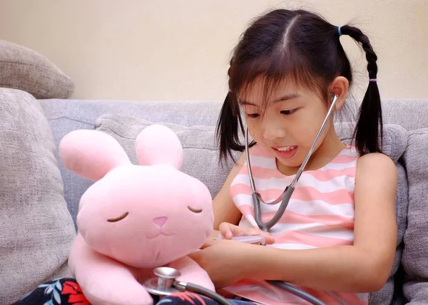 An Asian girl playing doctor with a stethoscope on her pink rabbit, smiling.