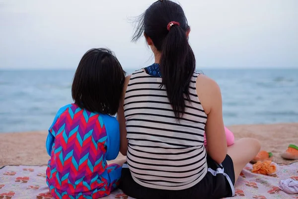 The back side of a mother and daughter sitting together by the beach, enjoying the view.
