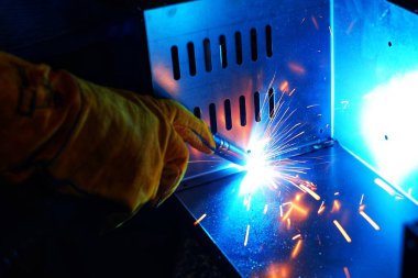 Pembe uzun kollu ceket giyen Hintli bir kadın, güvenlik maskesi ve deri eldivenler, bilgisayar sunucusu rafı için metal bir çanta kaynak yapıyor, kıvılcımlar.