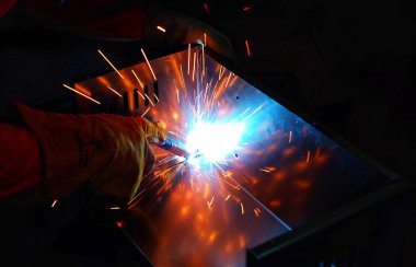 Pembe uzun kollu ceket giyen Hintli bir kadın, güvenlik maskesi ve deri eldivenler, bilgisayar sunucusu rafı için metal bir çanta kaynak yapıyor, kıvılcımlar.