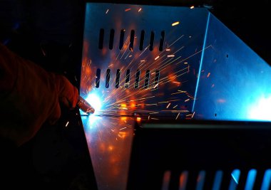 Pembe uzun kollu ceket giyen Hintli bir kadın, güvenlik maskesi ve deri eldivenler, bilgisayar sunucusu rafı için metal bir çanta kaynak yapıyor, kıvılcımlar.