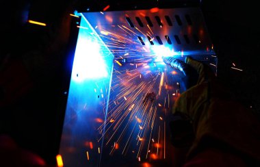 Pembe uzun kollu ceket giyen Hintli bir kadın, güvenlik maskesi ve deri eldivenler, bilgisayar sunucusu rafı için metal bir çanta kaynak yapıyor, kıvılcımlar.