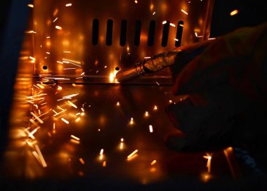 Pembe uzun kollu ceket giyen Hintli bir kadın, güvenlik maskesi ve deri eldivenler, bilgisayar sunucusu rafı için metal bir çanta kaynak yapıyor, kıvılcımlar.