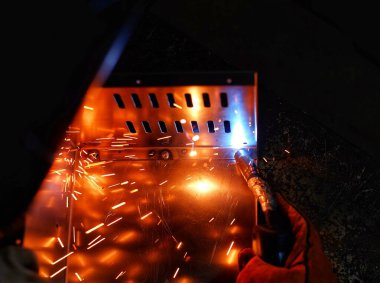 Pembe uzun kollu ceket giyen Hintli bir kadın, güvenlik maskesi ve deri eldivenler, bilgisayar sunucusu rafı için metal bir çanta kaynak yapıyor, kıvılcımlar.