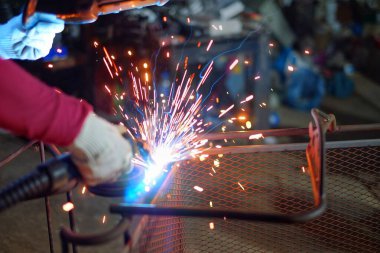 Pembe uzun kollu ceket giyen Hintli bir kadın, kaynak maskesiyle koruyucu eldivenler, kıvılcımlar saçan metal bir sandalye.