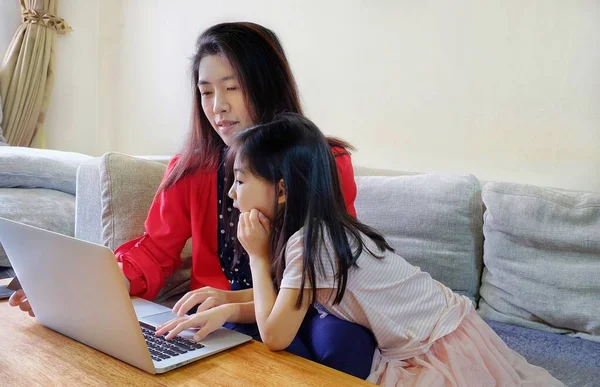 Çekici bir Asyalı kadın ve genç kızı kanepede oturuyor, internetten alışveriş yapıyor, bilgisayarını kullanıyor, eğleniyor ve birlikte vakit geçiriyorlar..