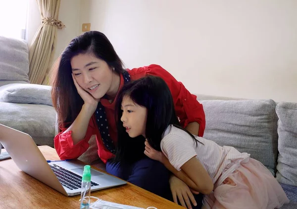 Çekici bir Asyalı kadın ve genç kızı kanepede oturuyor, internetten alışveriş yapıyor, bilgisayarını kullanıyor, eğleniyor ve birlikte vakit geçiriyorlar..