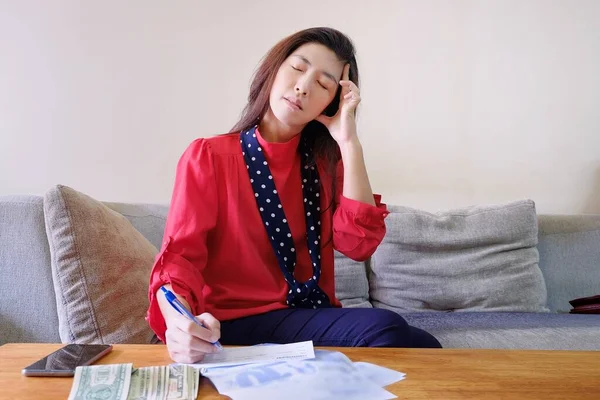 一位漂亮的亚洲女人 身穿红衫 坐在客厅的沙发上 背负着账单和债务 签着支票 — 图库照片