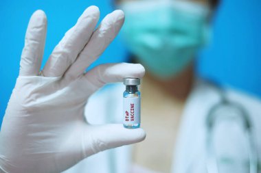 Klinikte maskeli ve beyaz lastik eldivenli bir kadın doktor elinde beyaz arka plan ve kırmızı harflerle bir şişe 1 doz DTaP ya da VIS aşısı tutuyor..