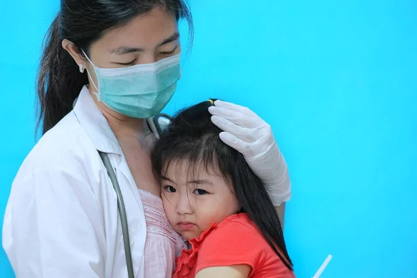 Aşı olmadan önce korkmuş ve gergin hisseden genç bir Asyalı kız, beyaz lastik eldivenli cerrahi maske takan doktoru tarafından teselli ediliyor..