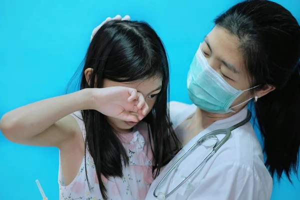 Aşı olmadan önce korkmuş ve gergin hisseden genç bir Asyalı kız, beyaz lastik eldivenli cerrahi maske takan doktoru tarafından teselli ediliyor..