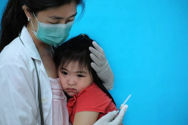 Aşı olmadan önce korkmuş ve gergin hisseden genç bir Asyalı kız, beyaz lastik eldivenli cerrahi maske takan doktoru tarafından teselli ediliyor..