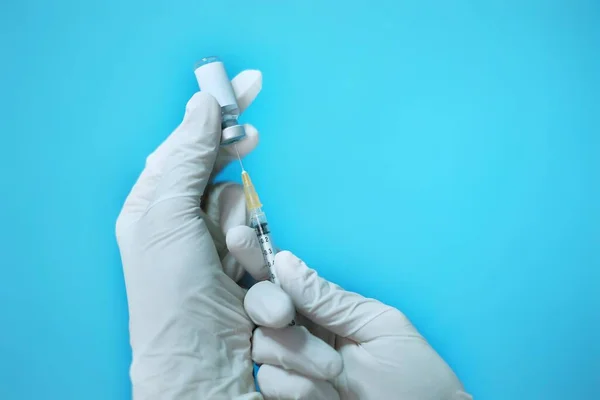 A female Asian physician with white rubber gloves and surgical mask at a clinic, inserting a needle and syringe on a clear glass bottle of vaccine with blank white label to inject it to a patient.