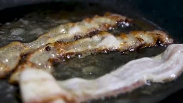クリスピーなベーコンスライスを油でフライパンに閉じます 豚肉のベーコンは熱い鍋でかき混ぜて喫煙します 朝食に不健康な食べ物を調理する — ストック動画