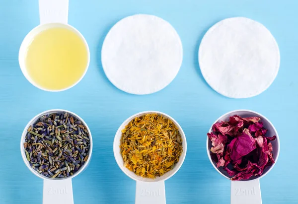 Scoop di plastica con olio d'oliva e varie erbe curative - calendula secca, lavanda e fiori di rosa canina. Aromaterapia, erboristeria e naturale concetto di cura della pelle. Vista dall'alto, copia spazio . — Foto Stock