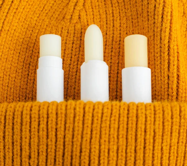 Three lip balms on the orange knitted background (pocket). Winter lip care sticks with beeswax, honey, panthenol and shea butter. Copy space.