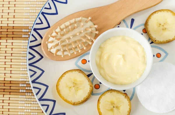 Diy banana mask (face cream) in the small white bowl, wooden hair brush, cotton pads and banana slices. Natural homemade hair treatment and zero waste concept. Top view, copy spac