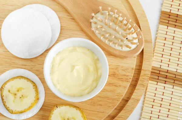 Diy banana mask (face cream) in the small white bowl, wooden hair brush, cotton pads and banana slices. Natural homemade hair treatment and zero waste concept. Top view, copy spac
