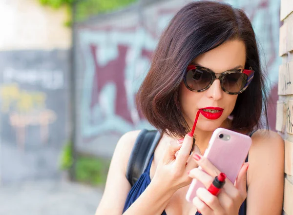 Seksi esmer kadın dudaklarını kırmızı rujla boyuyor. Bayan ayna olarak akıllı telefon kullanıyor; güneş gözlüklü çekici kız..