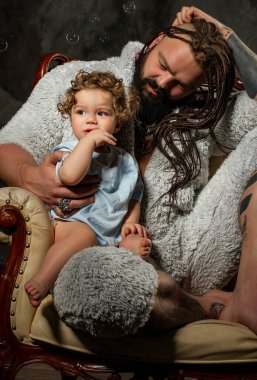 Kürk pijamalı baba çocuğa sevgiyle bakıyor. Sakallı ve dövmeli vahşi bir adam kollarında küçük kıvırcık saçlı bir çocuk tutuyor..
