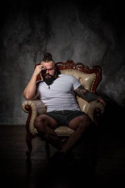 Sakallı ve dövmeli vahşi bir adam sandalyede oturuyor. Saç stili, tıraş edilmiş viski ve rastalı saç.