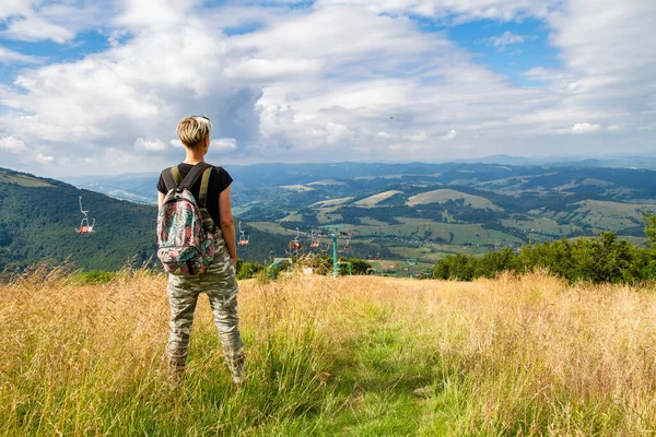 Kadın turist dağda duruyor, gökyüzüne, tepelere ve teleferiğe bakıyor.