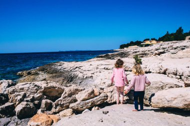 Deniz kenarında yürüyen küçük çocuklar kayalara tırmanıyor. Doğayı keşfeden iki kız kardeş. Kardeş konsepti. Mutlu çocukluk kavramı. Tatil zamanı. Yaz geldi. Kopya alanı olan aile tatilleri arka planı.