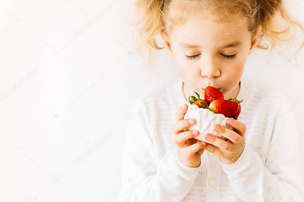 Lifestyle portrait of little blonde child eating homegrown strawberries on white background with place for text. Positive background. Slow living concept. Spring background. Joy of missing out.