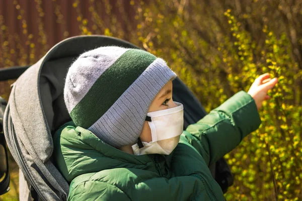 Dışarıdaki bir bebek arabasında tıbbi maskeli bir çocuk çiçek açan bitkilere uzanır.