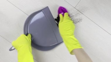 Sarı lastik eldivenli kadın, dağınık toprağı gri bir kepçeye süpürüyor.
