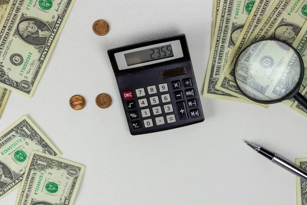 Banknotlar, madeni paralar, büyüteç ve kalemle beyaz bir masadaki hesap makinesiyle çalışıyor.