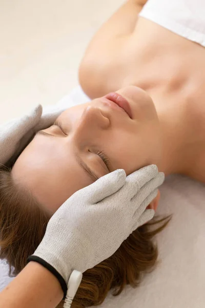 Kozmetik kliniğinde rahatlamış bir kadını yakından çek. Özel eldivenlerle mikro akım terapisi alıyor. Yüz bakımı.