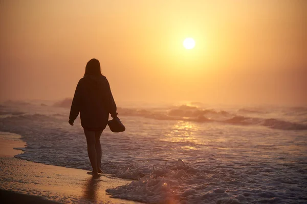 Bir kadın şafakta deniz kıyısında yürür, su sıçratır.