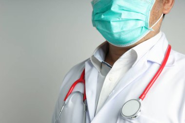 Yakın plan Asyalı bir adam doktor cübbesi giyiyor, tıbbi maske takıyor ve kırmızı bir steteskop kullanıyor. Beyaz bir arkaplanda izole edilmiş ve sol tarafında kopya alanı var. Kavram koruma virüsü.