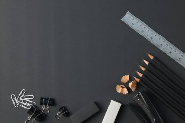 Ofis kırtasiyesi üst görünümü. Çalışma alanı kalemler, cetvel, silgi, ataç, zımba, siyah masa düz uzanıyordu..