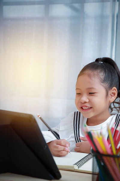 亚洲女孩很高兴在家网上做作业 在Coronavirus期间为儿童提供远程教育 她高兴地笑着 一边写着一本书 一边打电话给老师 — 图库照片