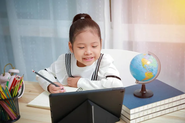 亚洲孩子在家里上网学习 在Coronavirus期间为儿童提供远程教育 她通过视频电话愉快地写着答案和微笑与老师交流 — 图库照片