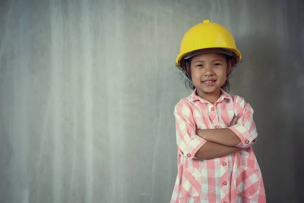 İnşaat kaskı takan küçük kız kollarını kavuşturdu. arkaplan gri çimento kopyalama alanı.